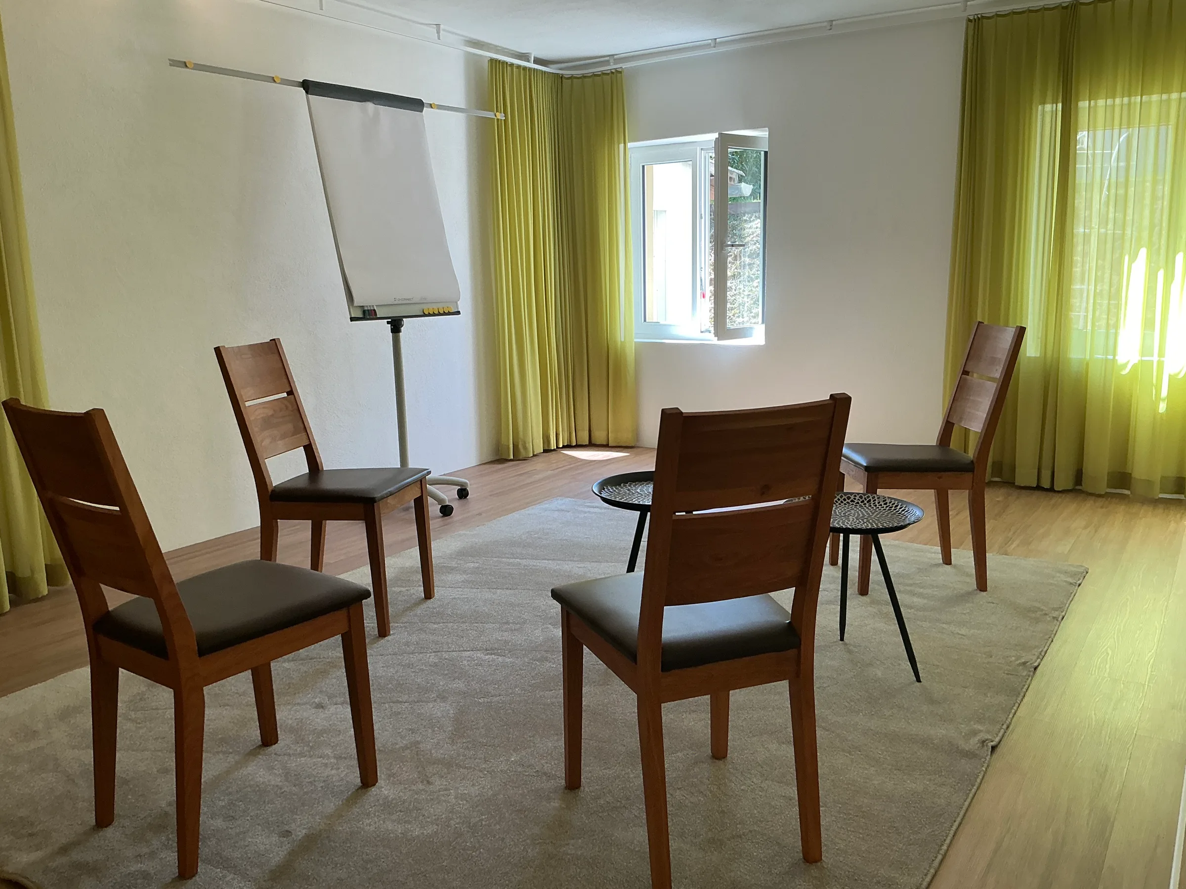 Praxisraum mit vier Holzstühlen im Halbkreis, Flipchart und limettengrünen Vorhängen